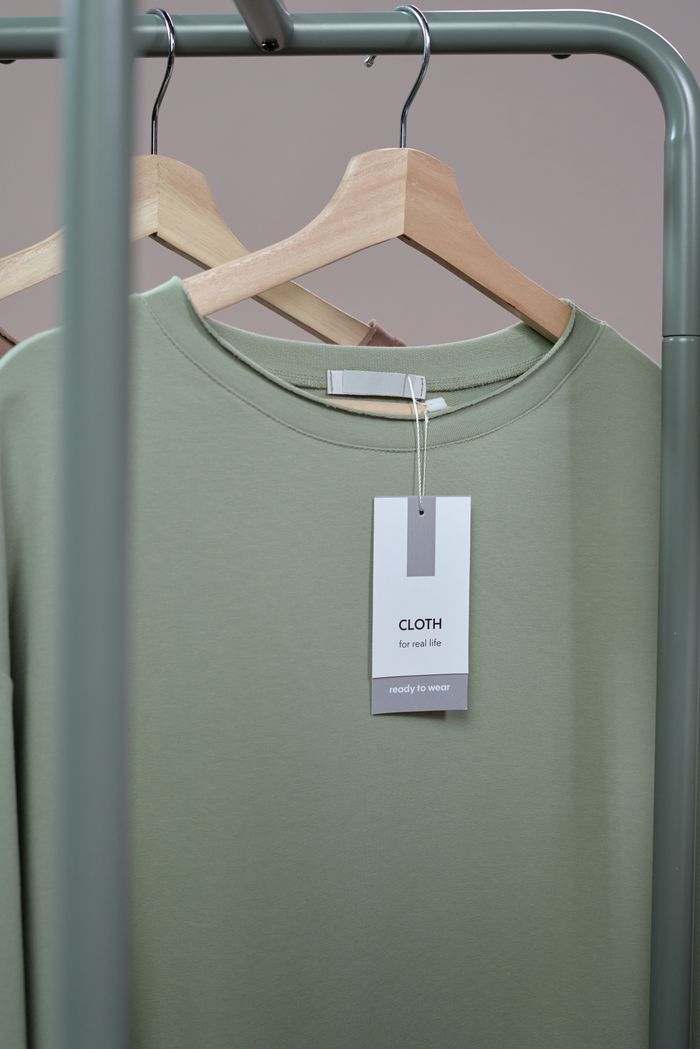 A simple green t-shirt with a tag hanging on a wooden hanger against a neutral background.